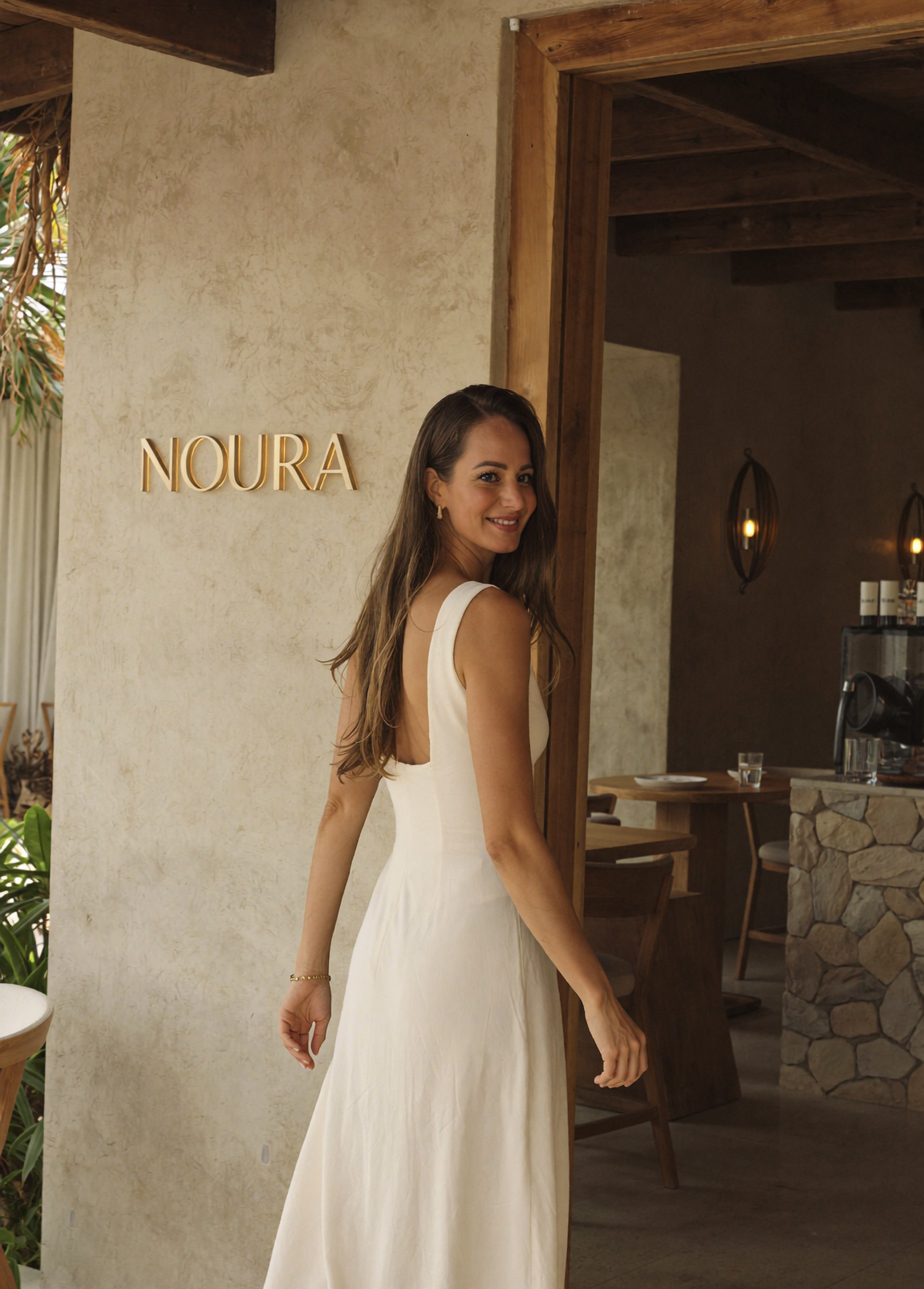 Woman in a cream backless dress smiling over her shoulder outside the cafe entrance, beside gold NOURA lettering on a sandy beige wall