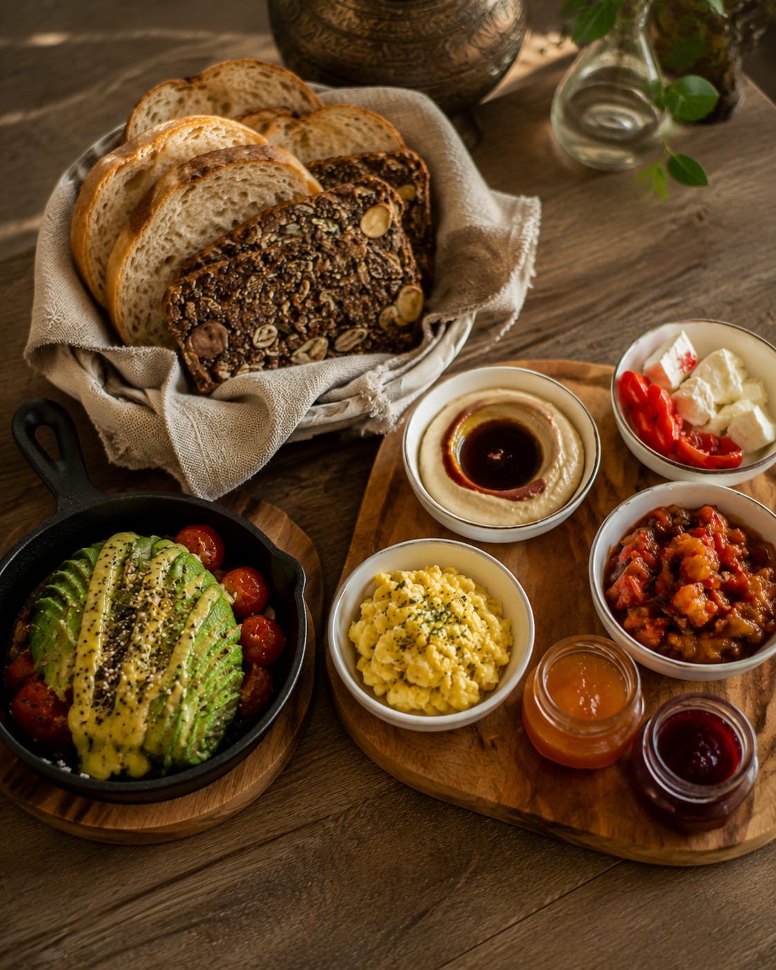 Brunch spread with bread basket, avocado and roasted tomatoes in a cast-iron skillet, and small bowls of hummus, eggs, cheese and jams on a wooden board