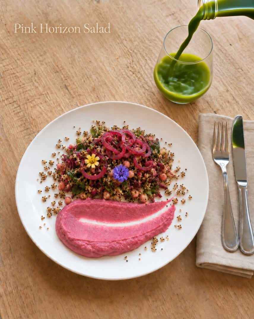 Top-down view of The Pink Horizon salad plated over a swipe of pink beet puree beside a glass of green juice